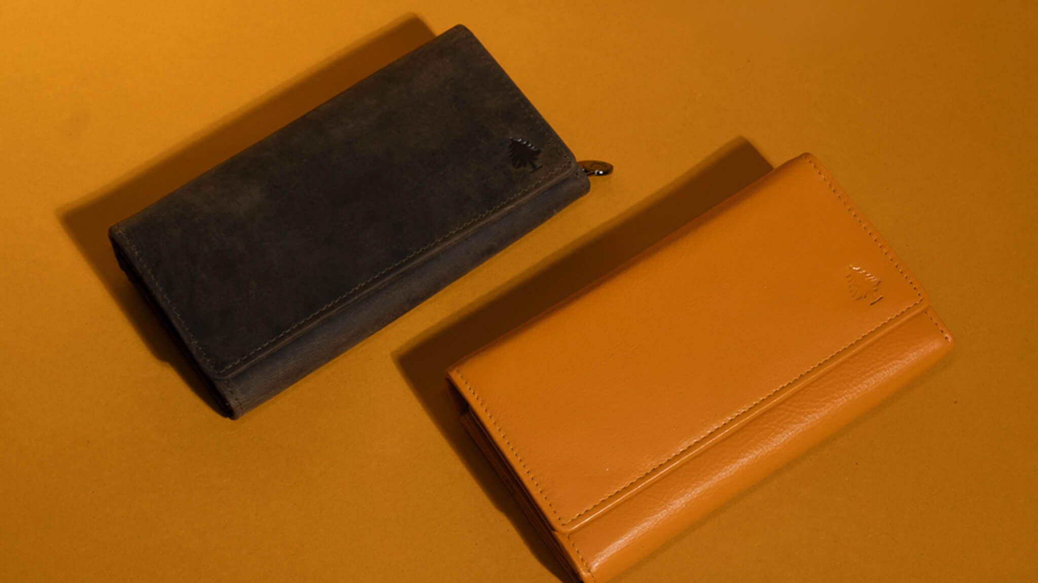 Zwei lange Leder-Geldbörsen auf warmgelbem Hintergrund – eine olivgrüne Wildleder-Geldbörse und eine glatte senfgelbe Leder-Geldbörse im minimalistischen Design